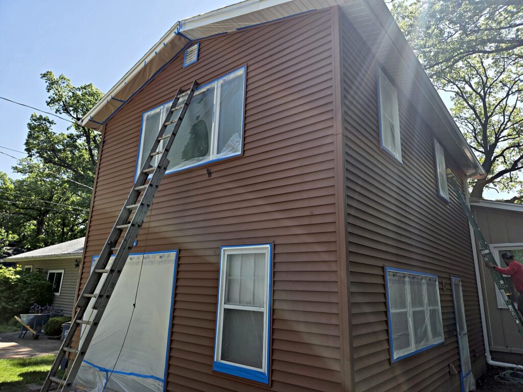 Two-story house with brown siding, a ladder and windows masked with plastic in West Bend